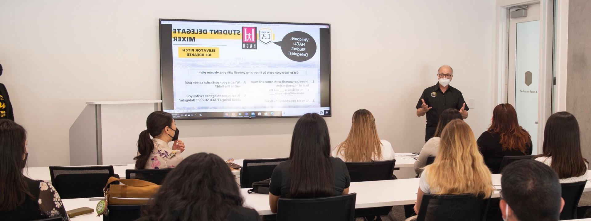 Person wearing a mask speaking in front of a class, standing next to a smart screen.